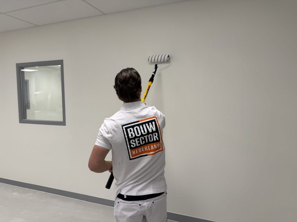 Schilder in witte werkkleding zet met de roller een baan verf op een lichte muur bij een binnenraam