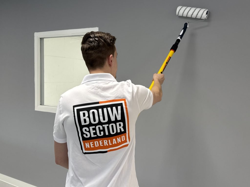 Schilder met Bouwsector-logo op de rug rolt grijze verf op een muur naast een wit raamkozijn