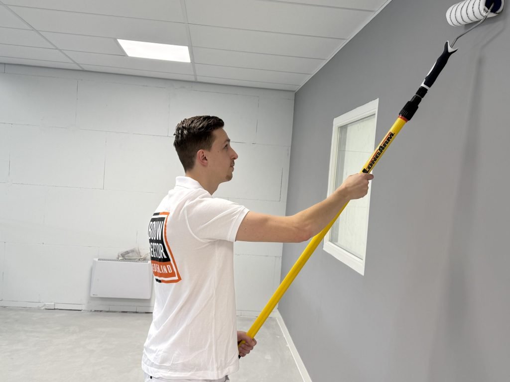 Schilder in wit poloshirt rolt een grijze muur naast een raam met de roller aan een lange steel