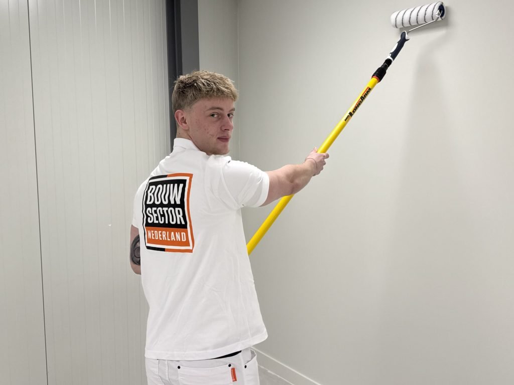 Schilder kijkt over zijn schouder terwijl hij een lichte muur in een smalle ruimte sauzt