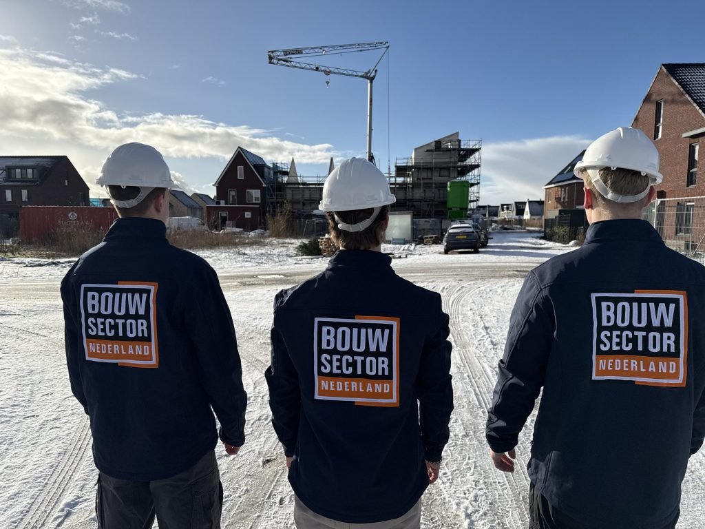 Drie vakmensen van Bouwsector Nederland met witte helm op een besneeuwde weg tussen bakstenen woningen