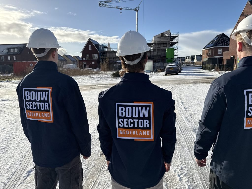 Drie vakmensen van Bouwsector Nederland met witte helm en donkere jas kijken naar een straat met nieuwbouw
