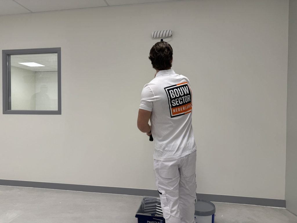 Schilder van Bouwsector Nederland rolt een witte wand in een lege ruimte, met verfbak en emmer op de vloer
